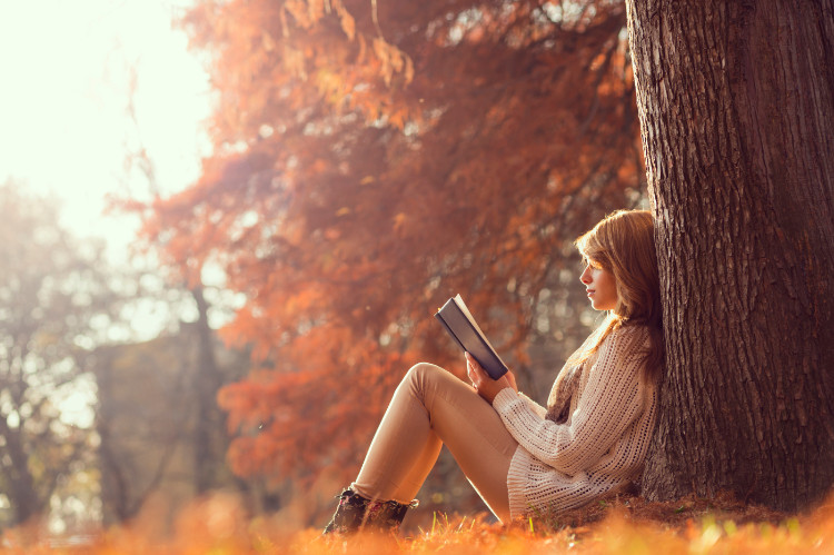 donna seduta nella foresta, appoggiata a un albero, che legge un libro in autunno