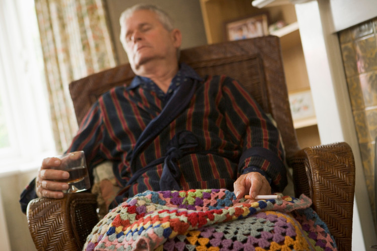 un nonno addormentato su una sedia coperta da una coperta, con un bicchiere di alcol e una sigaretta in mano