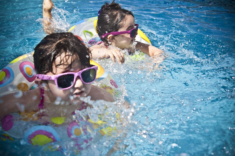 Due bambini nuotano nell'acqua, indossano occhiali da sole con montatura rosa e sono su biciclette galleggianti.