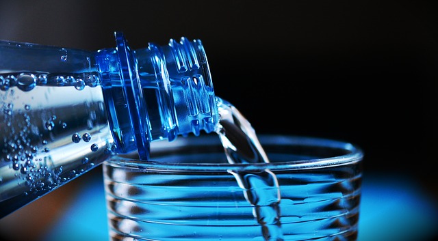 L'acqua viene versata dalla bottiglia nel bicchiere. Sono visibili solo il collo della bottiglia e la parte superiore del bicchiere. Lo sfondo è scuro e blu.