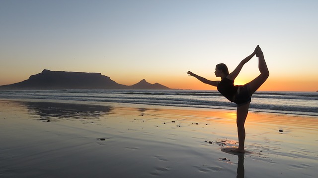 Una donna sta praticando yoga sulla spiaggia. Il sole è tramontato, ma c'è ancora luce. Lei sta in piedi su una gamba sola. C'è la bassa marea, la sabbia è sotto un sottile strato d'acqua.