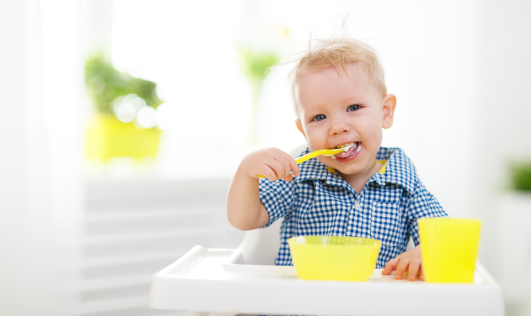 Un bambino piccolo, seduto su un seggiolino, si nutre da solo. Cucchiaio, tazza e ciotola gialli.