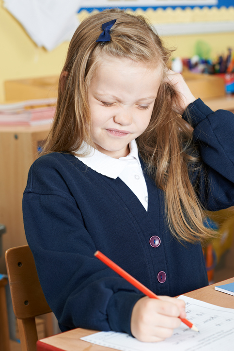 Una bambina seduta a scuola con un prurito alla testa, probabilmente pidocchi nei capelli, capelli biondi, seduta a scrivere