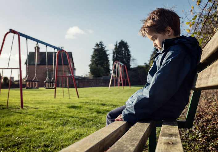 ragazzo seduto da solo su una panchina, triste, che prova emozioni negative, parco giochi, erba, altalena