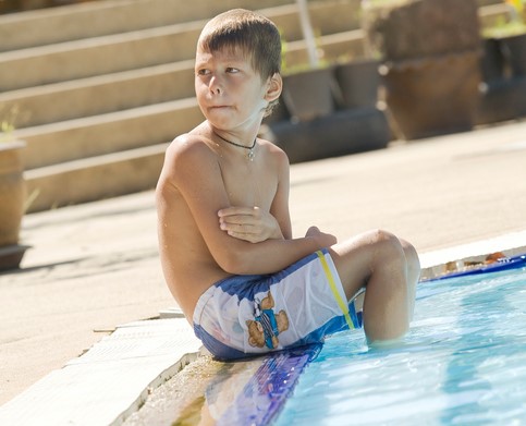 Il ragazzo è seduto vicino alla piscina, con i piedi nell'acqua, e ha freddo.